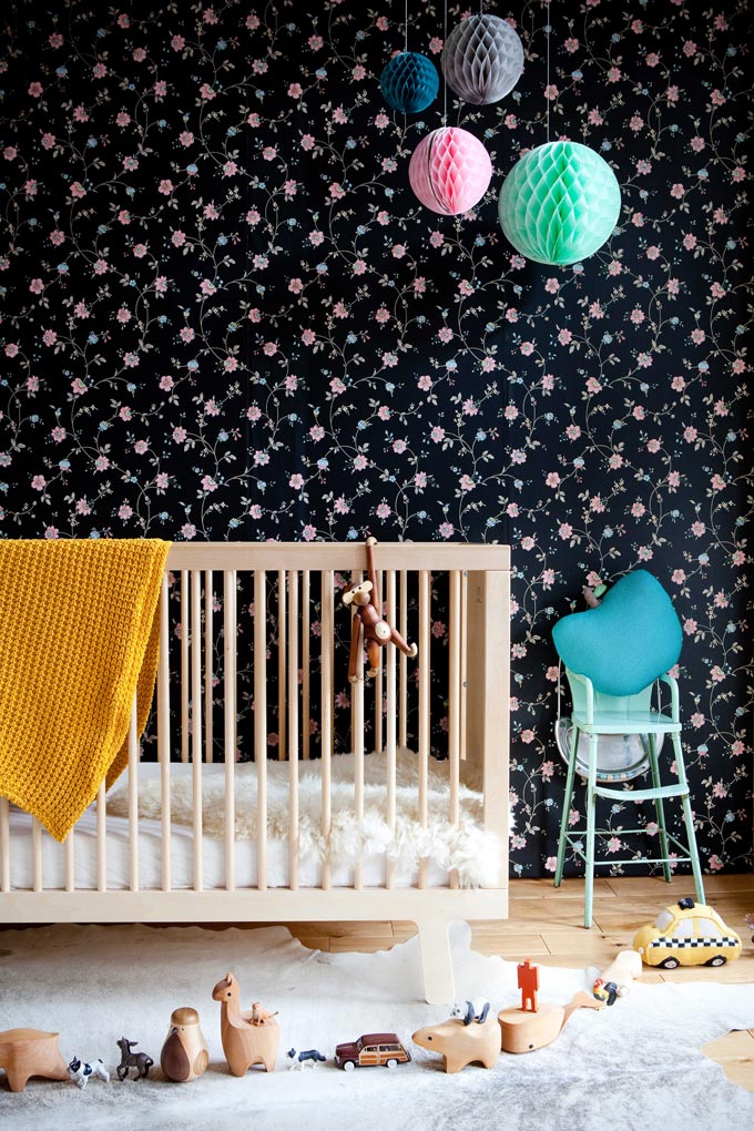 A dark blue wallpaper with a flower pattern looking amazing in a nursery room with a wooden crib and pops of color. Image by Cuckooland.