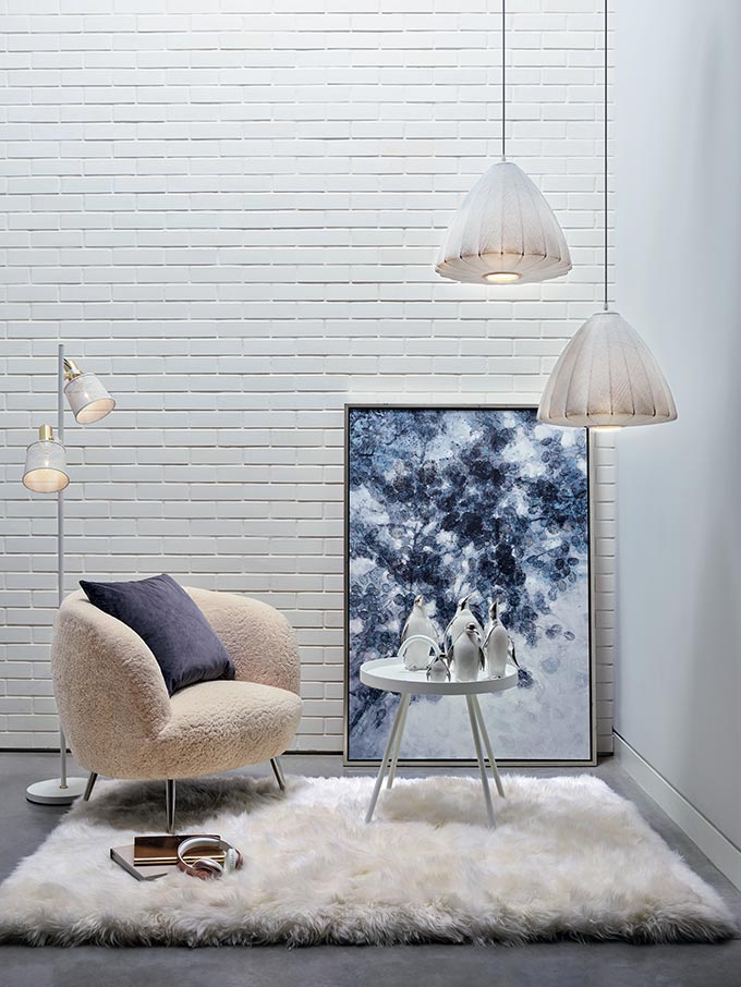 A stylish set composed of an armchair in shearling over a sheeprug, with an oversize artwork against a white brick accent wall and white pendant lights hanging over a small round side table. Image by Homesense.