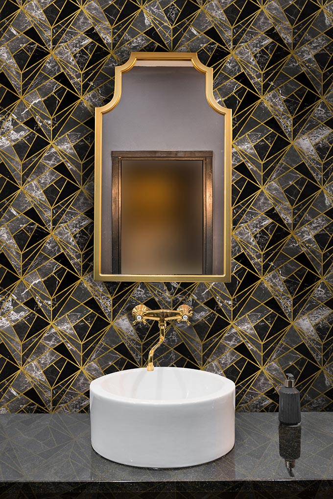 A beautiful geometric pattern wallpaper looks stunning in this bathroom with a vintage looking brass framed mirror over a round washbasin. Image by Mind the Gap.