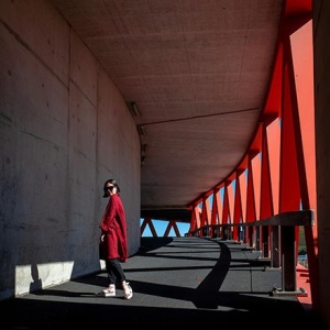 A woman in a red coat standing outdoors under the roof canopy of a concrete building with a steel structure on the side. Image by Artem Gavrysh on Unsplash.