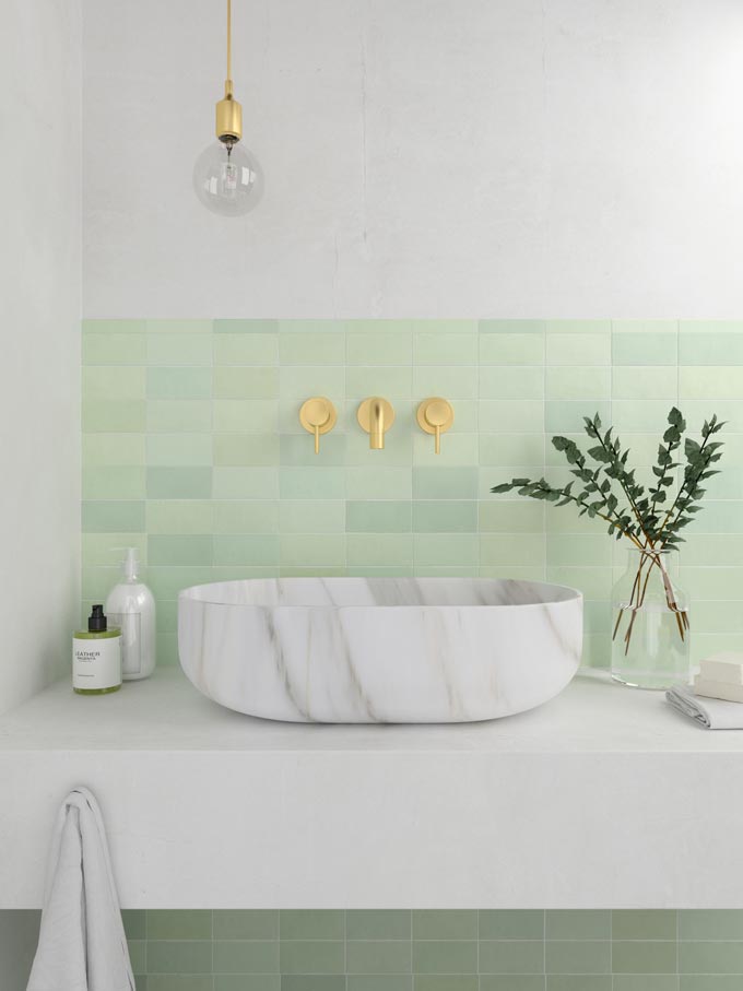 A gorgeous bathroom with a marble looking washbasin and a tile backsplash with soft green tiles and brass fixtures. Image by WOW Design.