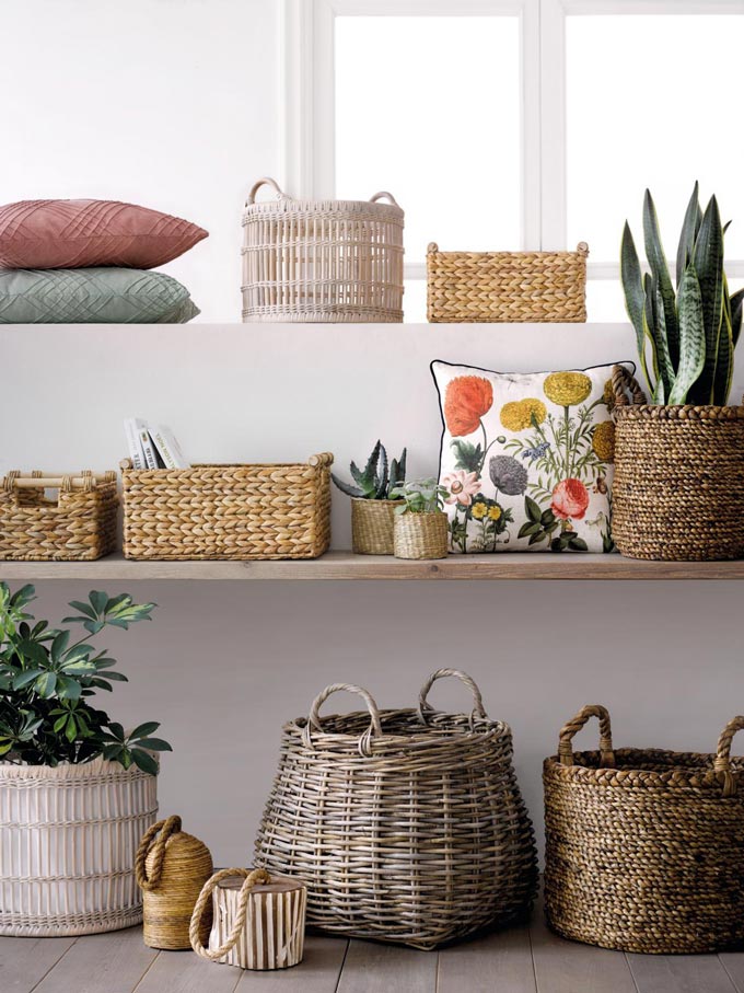 Open shelves stacked with rattan baskets, plants, cushion and decorative pillows. It has a bohemian flair to it but in a very refined way. Image by El Corte Inglés Decoración.