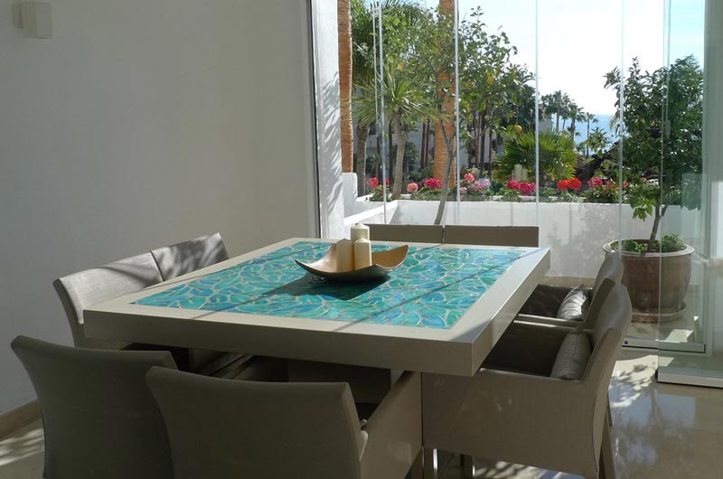 A contemporary dining space next to large window doors with a dining table whose table top has been laid with turquoise leafy pattern handmade tiles. Image by G. Vega. 