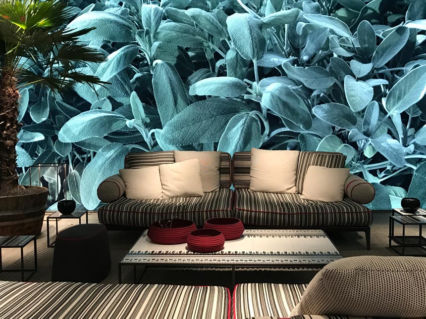 View of a stunning living space with a magnified blue green leafy accent wall as a background to a pattern print sofa. This image is from the stand of Poliform at the imm Cologne 2019 fair.