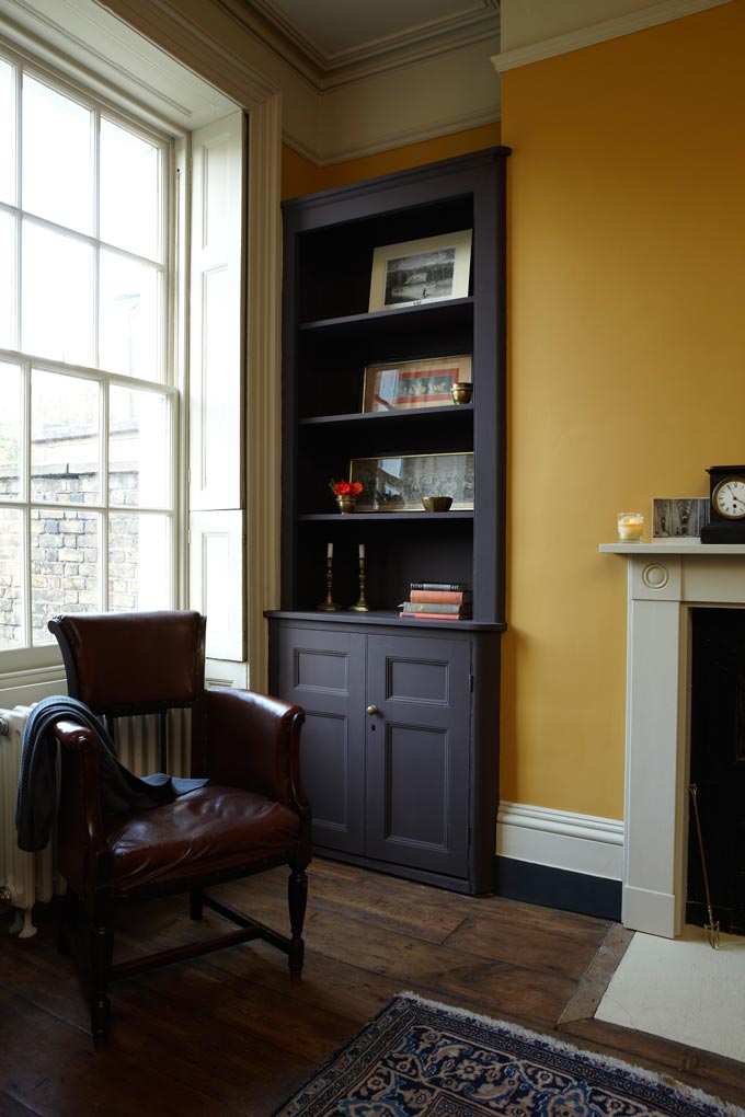 What a cozy vignette by a window with a dark built in bookcase, a fireplace and bold ochre yellow wall to contrast those rustic but sophisticated tones. Image by Farrow and Ball.