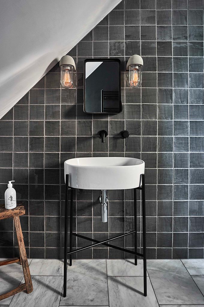 Love the white grout between the grey square tiles in this bathroom with a white washbasin on a black metal stand. The bathroom looks complete with the two sconces next to the mirror. Image by Garden Trading.