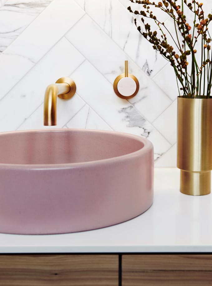 Detail view of a white marble looking tile backsplash in a herringbone pattern, golden faucet and pink washbasin. Image: Norsu interiors