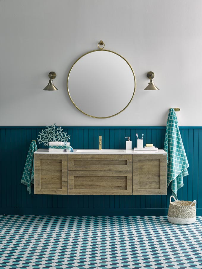 A contemporary bathroom where teal reigns in the wainscotting and flooring. A brassy round mirror is paired nicely with two brassy sconces on each side of it. I like the wall mounted wooden tone vanity furniture that makes everything feel more airy. Image by El Corte Inglés Decoración.