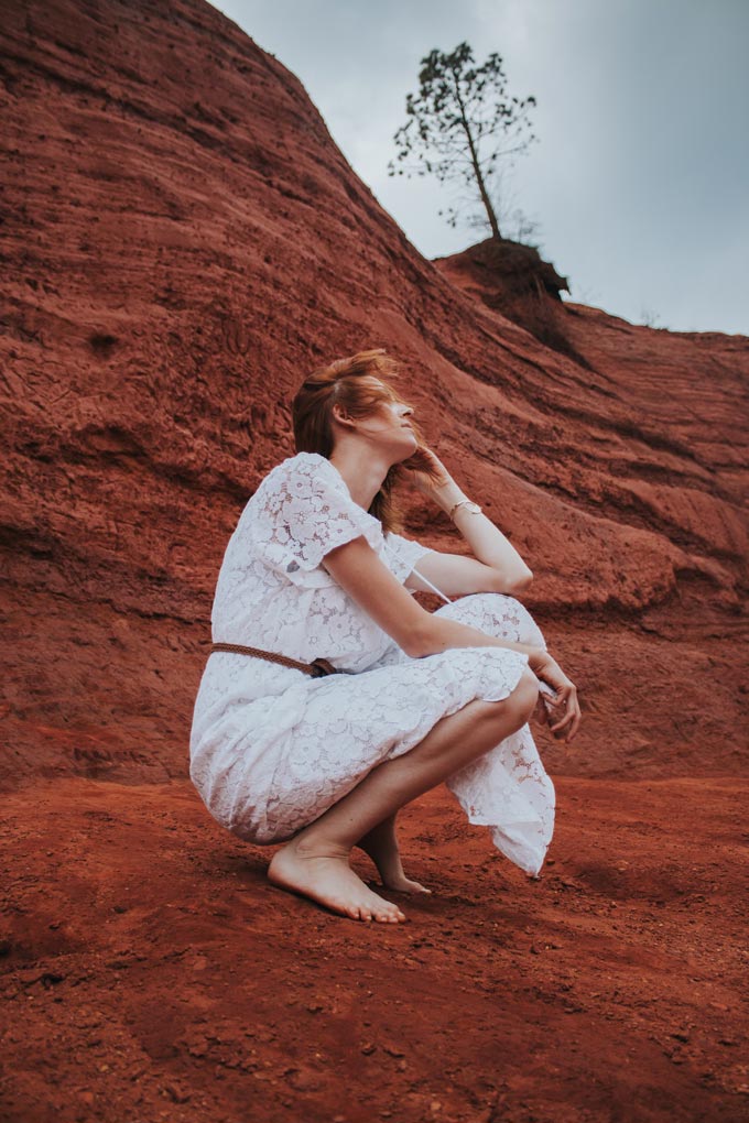 A barefoot red haired woman wearing a summer dress sitting on a red brown rocky mountain side.