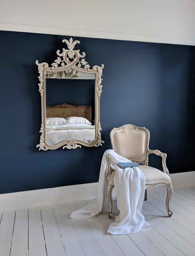 A rectangular mirror with heavy ornamentation against a dark blue wall, next to an off white armchair. Image by The French Bedroom Co.