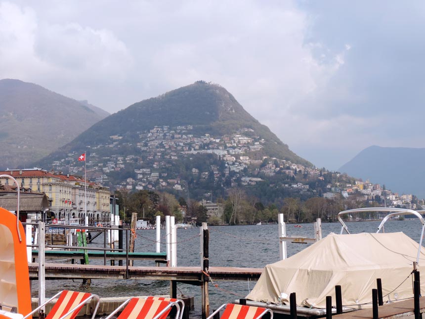 Partial view of Lugano in the background and the lake with some docked paddle boats on the shore.