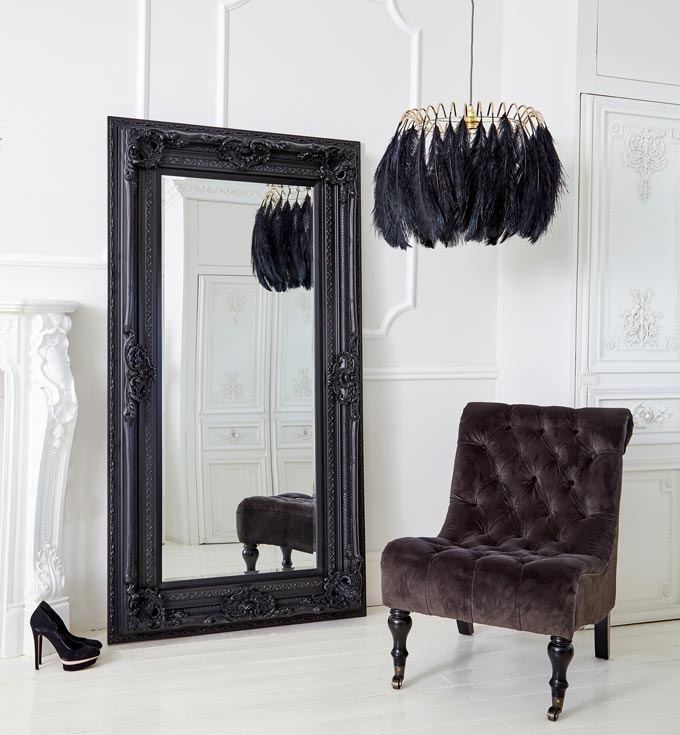 A double black framed French style oversize mirror against a white paneled wall along with a statement pendant light made with black feathers and a black velvet armless chair. Image by The French Bedroom Co.
