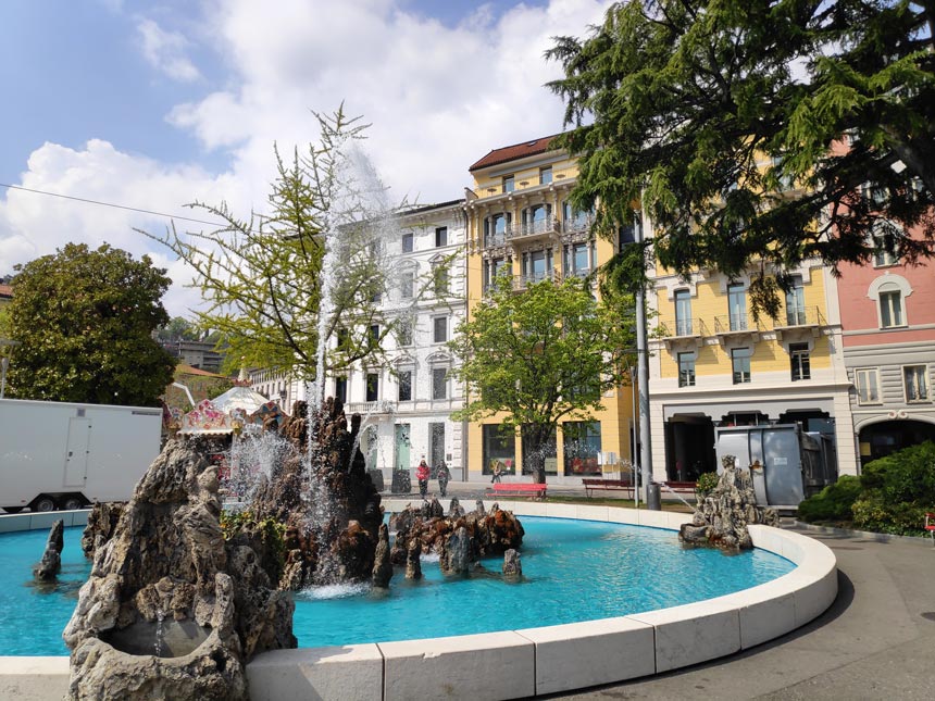 A water fountain in the foreground and old buildings in the background from the old town of Lugano.