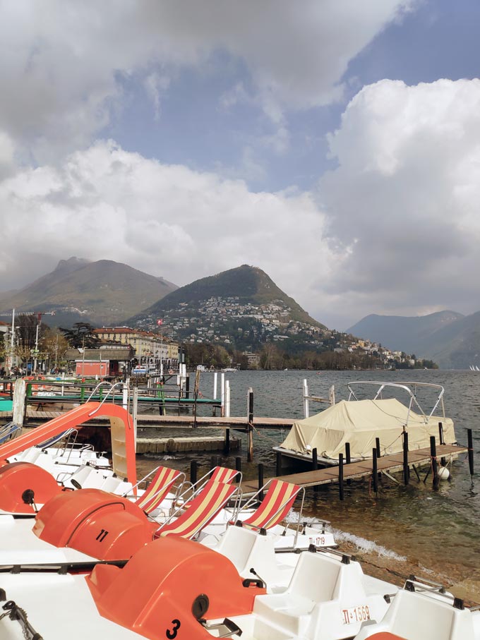 Partial view of Lugano in the background and the lake with some docked paddle boats on the shore.