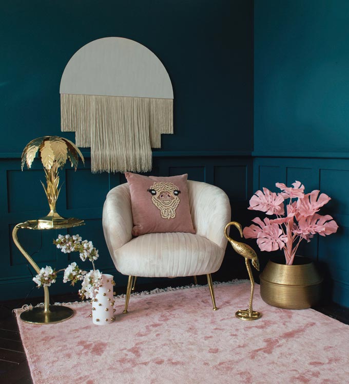 These teal hue wall colors are the perfect background for a curvy off white armchair sitting atop a blush pink area rug and with a half circle mirror decorated with a hanging long fringe. Image by Audenza.