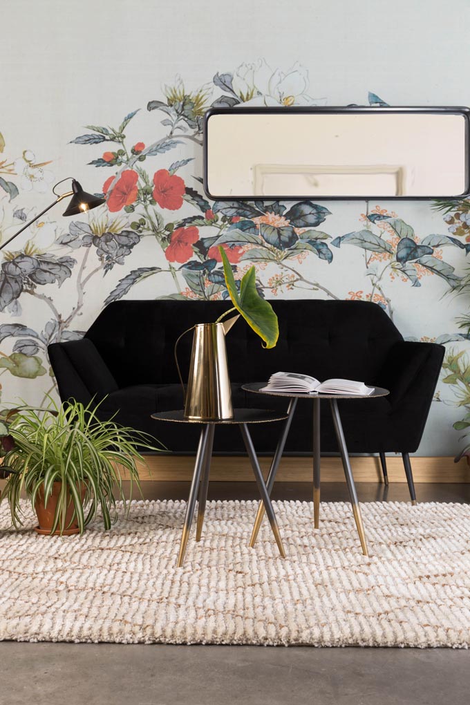 A rectangular mirror with rounded edges is placed horizontally over a black sofa, adding depth against a flower wallpaper mural. Image by Cuckooland.
