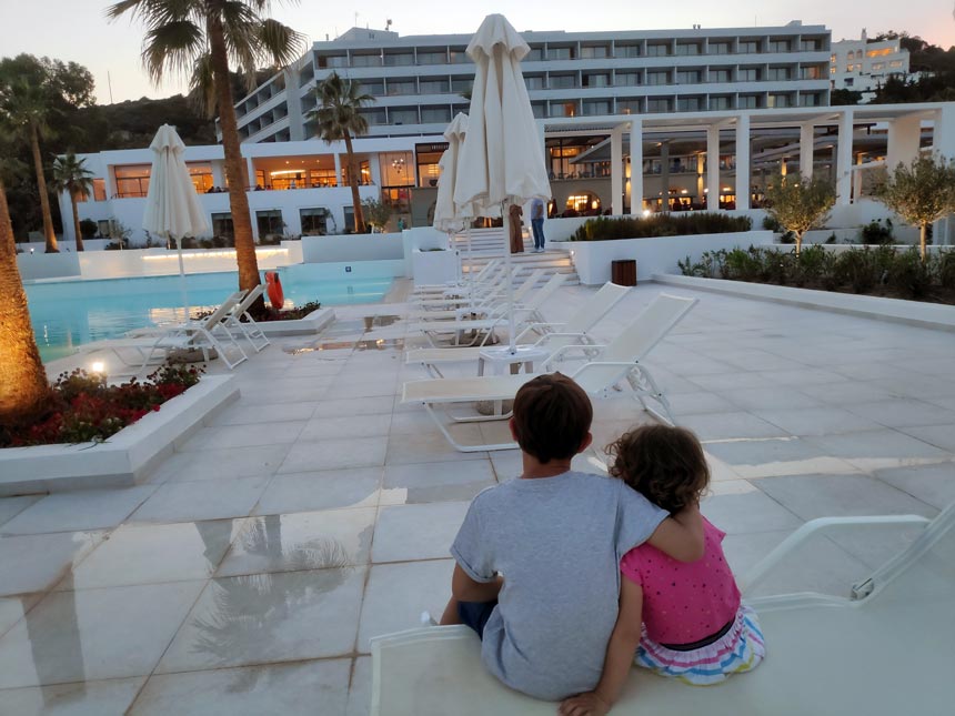 My two children sitting on a sunbed by the main pool of Grecotel Lux Me Rhodos looking at the main building.