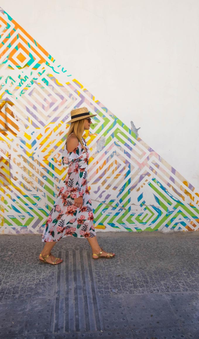 A woman wearing a flower print dress is walking by a wall with a colorful pattern to it.