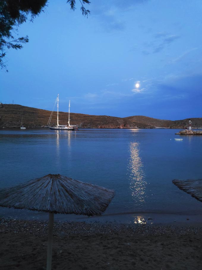 The moon peaking through the clouds casting a glare over the blue waters and the sailing boat with its lights on frame this idyllic view from the beach of Achladi in Syros.