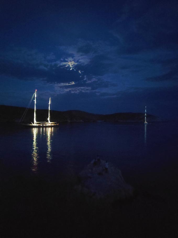 Two sailing boats in a cove while the moon is covered with clouds, barely peaking.