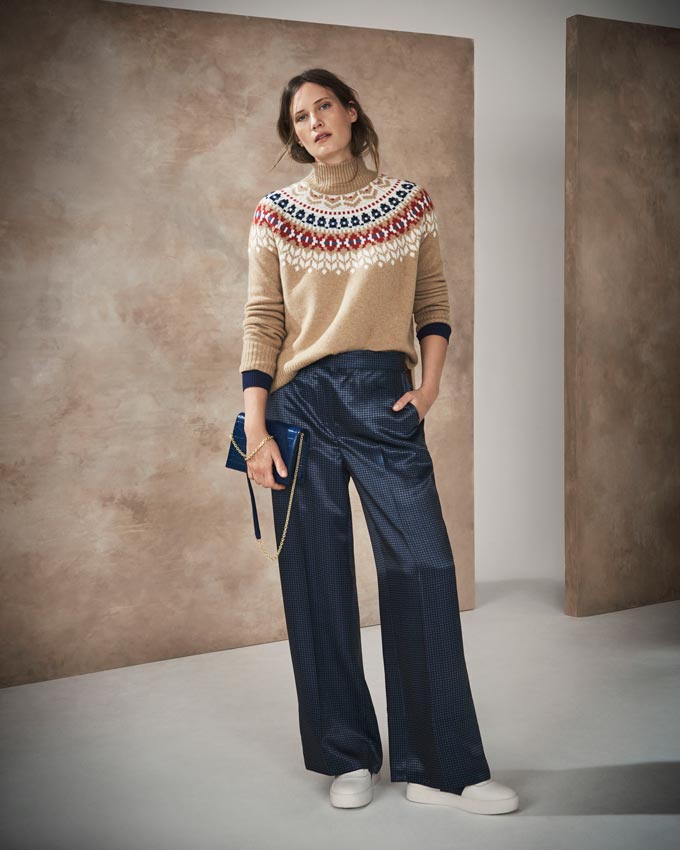 Fall fashion ideas: I like this combo of a beige with a pattern around the neck knit over some loose straight dark blue shiny pants paired with a handbag of the same color and white trainers. Image by Marks&Spencer.