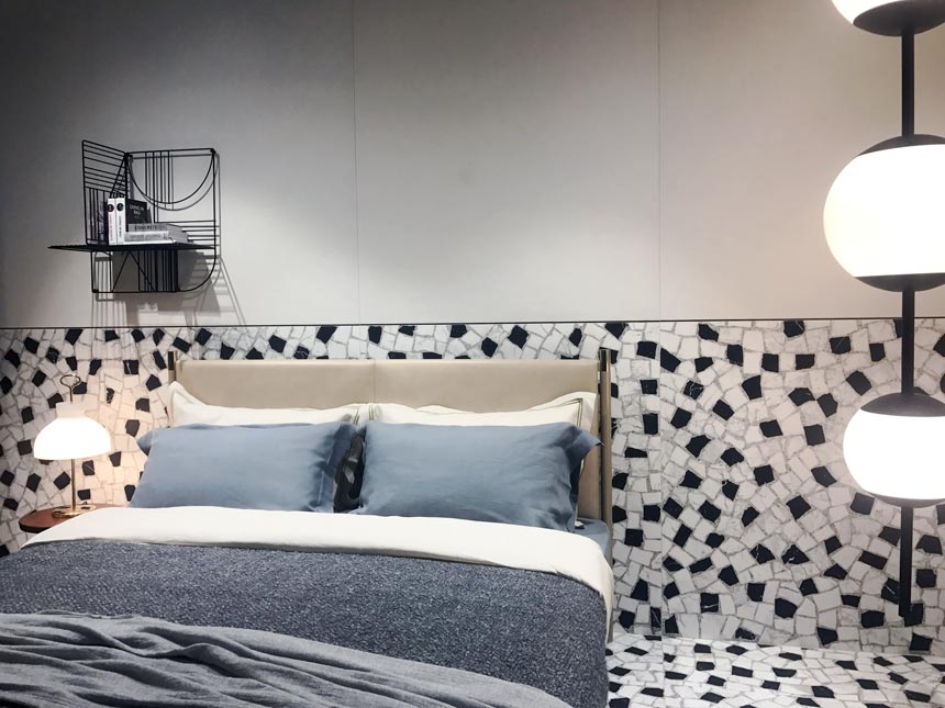 Terrazzo looking tiles extending from the floor up to half way up the wall to make an accent behind a neutral contemporary bed. All that as seen at Ragno's stand at Cersaie 2019. 