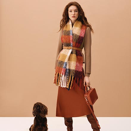 A pasmina worn over a camel coat and a burnt orange skirt and slouchy boots. Note the colors both that cute dog and woman sport so well. Image via Hobbs.