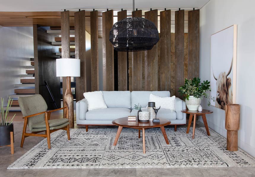 A chic Scandi-boho inspired living room with a soft grey sofa, a large black rattan pendant light, a patterned in neutral colors area and lots of wooden surfaces. Image via OZ Design Furniture.