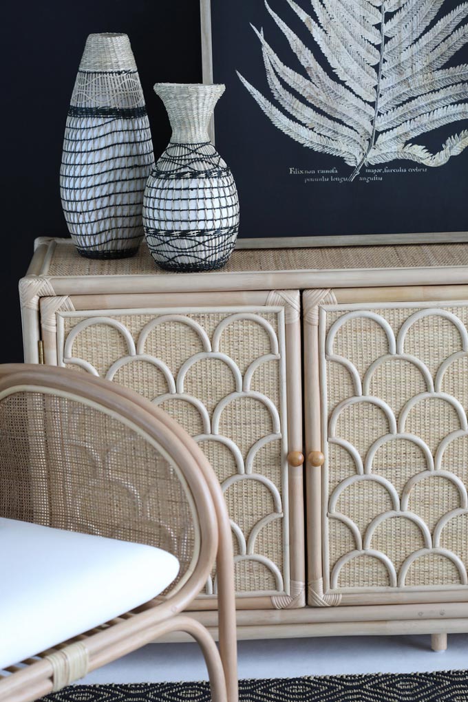 Detail view of a vignette featuring a beautiful credenza made of rattan and cane, decorated with some patterned bases.Alongside is a handmade rattan chair. The navy blue wall color behind helps them pop up. Image via OZ Design Furniture.