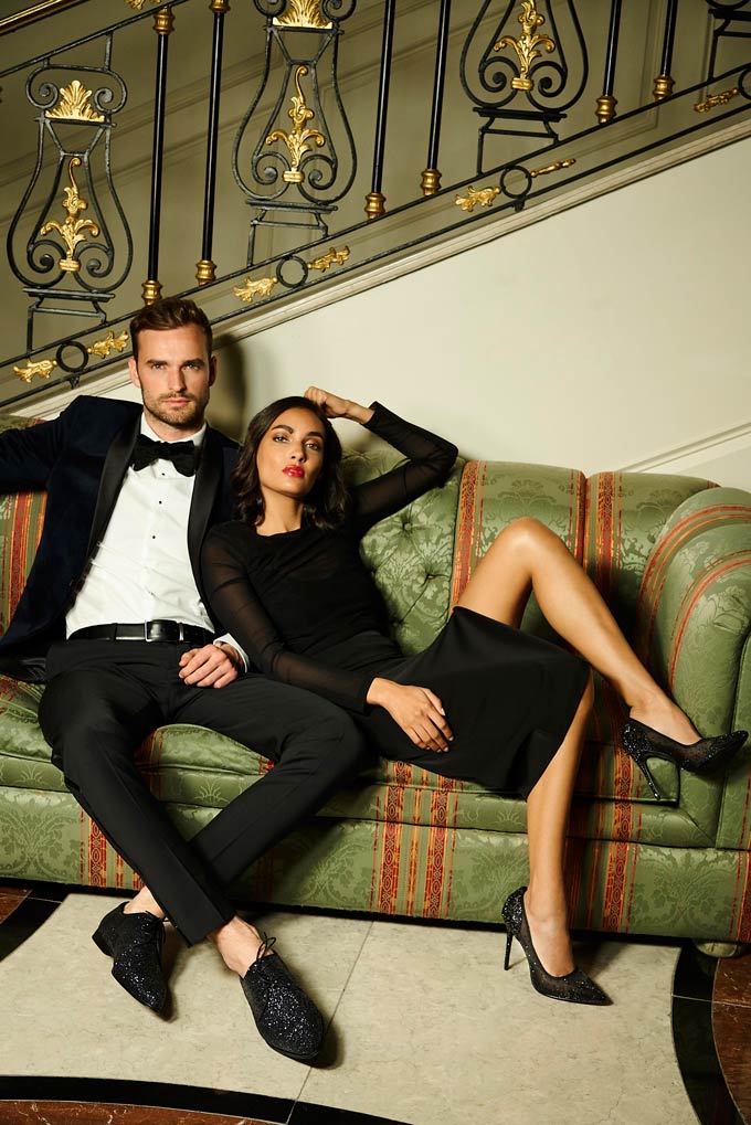 A handsome couple dressed up in holiday outfits. He's wearing a black suite, she's wearing a velvet dress with shears and black glittering high pumps. Image via Dune.