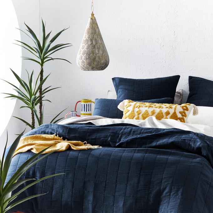 A white bedroom with a bed covered in navy blue bedding and pops of mustard hue accents. Image via Adairs.