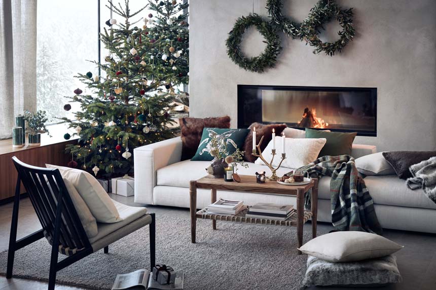 A beautiful contemporary living space with large windows, a Mid-century inspired armchair, an off white sofa in front of a modern fireplace and lots of Christmas decor on the coffee table and Christmas tree. Image via H&M homestores.