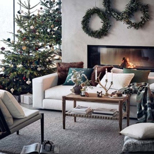 A beautiful contemporary living space with large windows, a Mid-century inspired armchair, an off white sofa in front of a modern fireplace and lots of Christmas decor on the coffee table and Christmas tree. Image via H&M homestores.