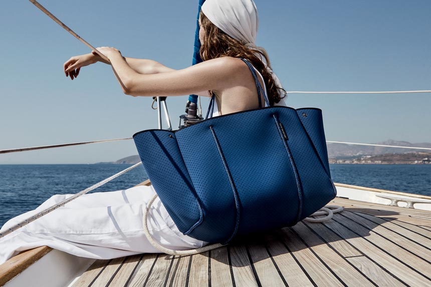 A woman on board a sailing boat carrying a large classic blue tote bag sitting on the deck and gazing at the sea. Image via State of Escape.