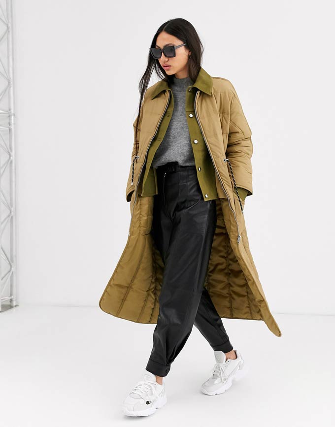 A mustard tone overcoat paired with a grey top, black faux leather wide leg pants, and white trainers. Image by ASOS.