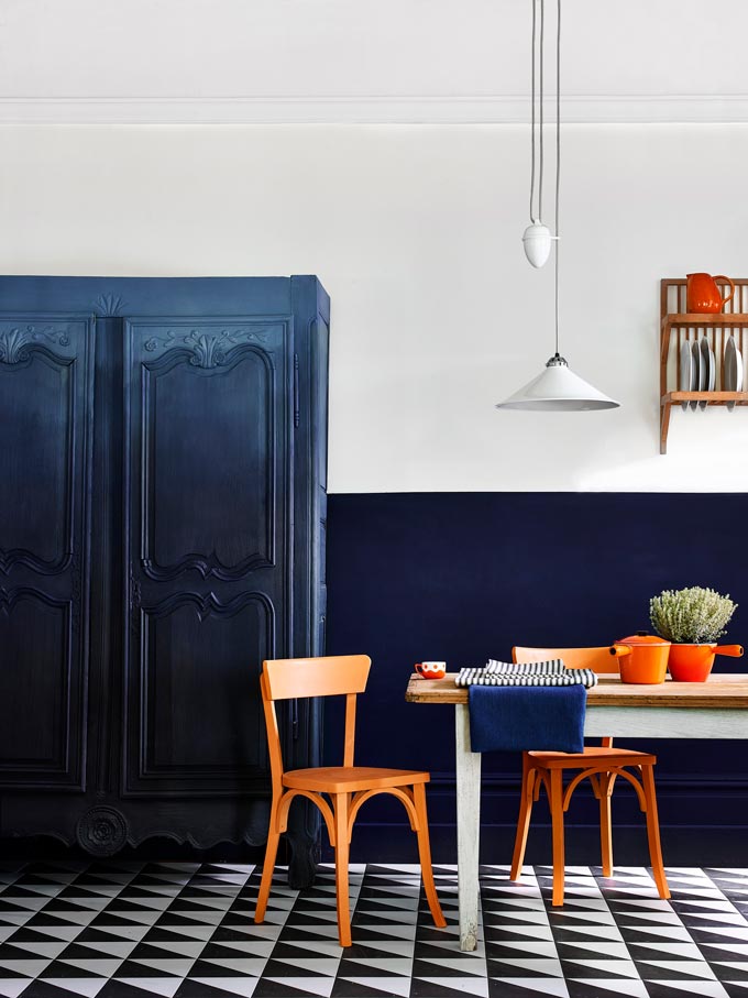 A color blocked kitchen with a blue and white wall, a pattern tile flooring, an ombre effect blue vintage armoire next to a dining set with orange chairs and decor. Image via Annie Sloan.
