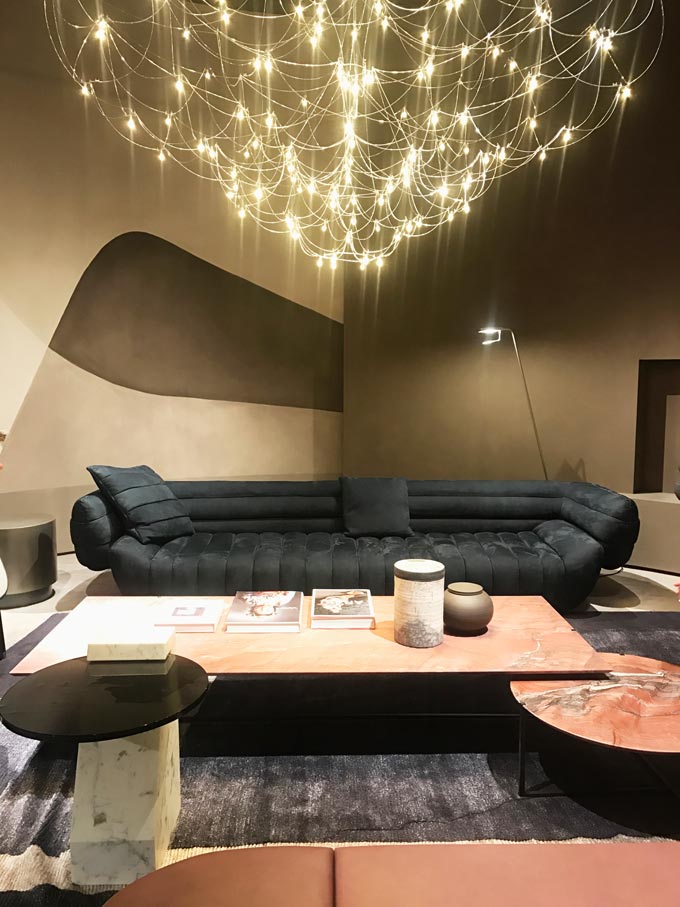 A contemporary installation with a dark grey sofa and pink marble coffee tables and a gorgeous huge delicate and modern chandelier as seen at Baxter.