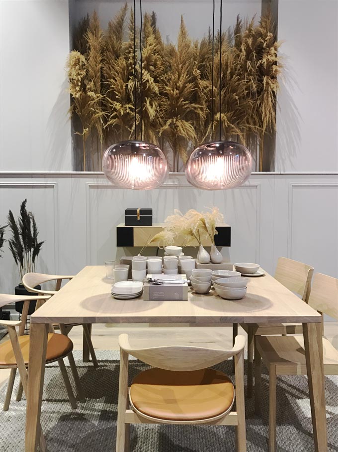 From Bolia. A dining set in light wood and two pendants lights. Pampas grass is used as decor in the background.