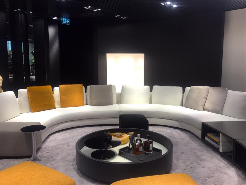A big contemporary sitting lounge in a circular arrangement with a round black coffee table in the middle. Everything has a vibe from the 70's. As seen at Minotti.