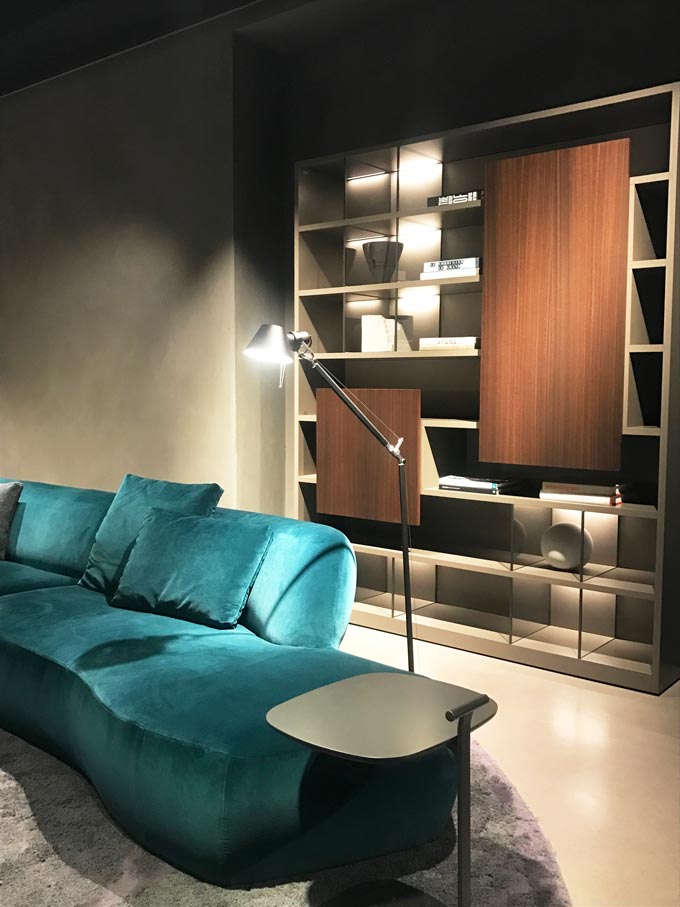 A teal velvet sofa in a contemporary setting in front of a dark stained wooden bookcase from Molteni at the imm Cologne 2020.