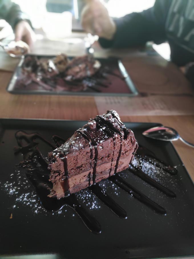 A slice of chocolate cake in the foreground and two people eating another in the background. The image was captured using the portrait mode of a smartphone camera.