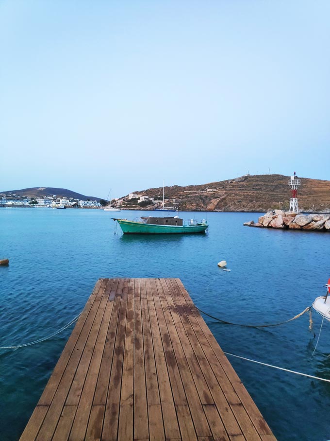 An old green fishing boat within a cove a bit far off from a wooden narrow dock. A beacon in the background and the wooden dock in the foreground. A smartphone image by Velvet.