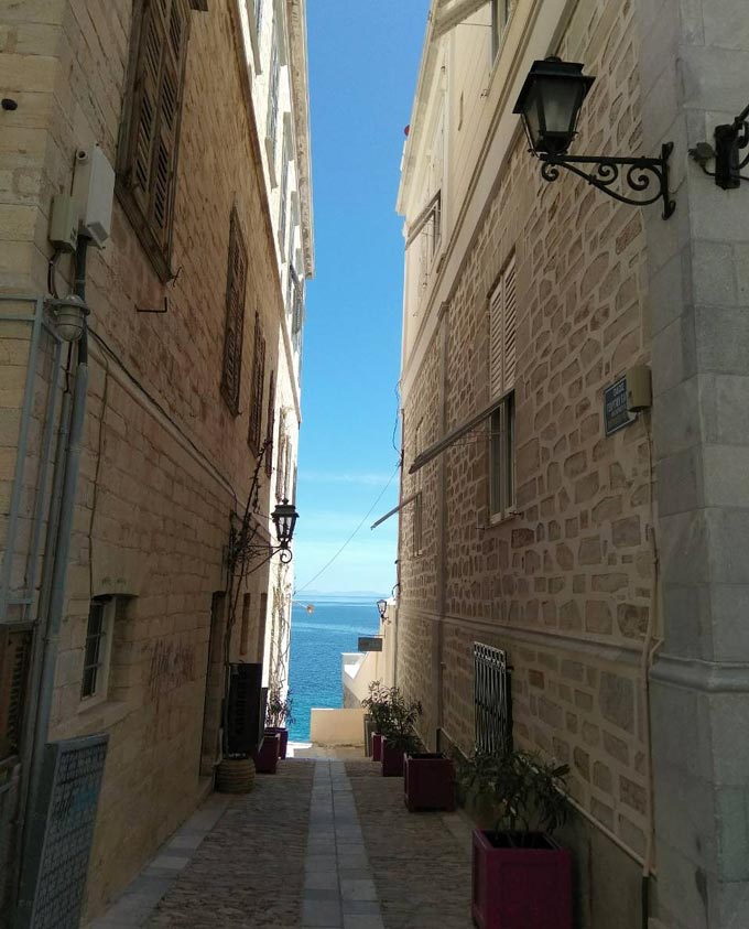 A narrow stone paved alley lending view to the sea between two old stone buildings in Hermoupolis, Syros.