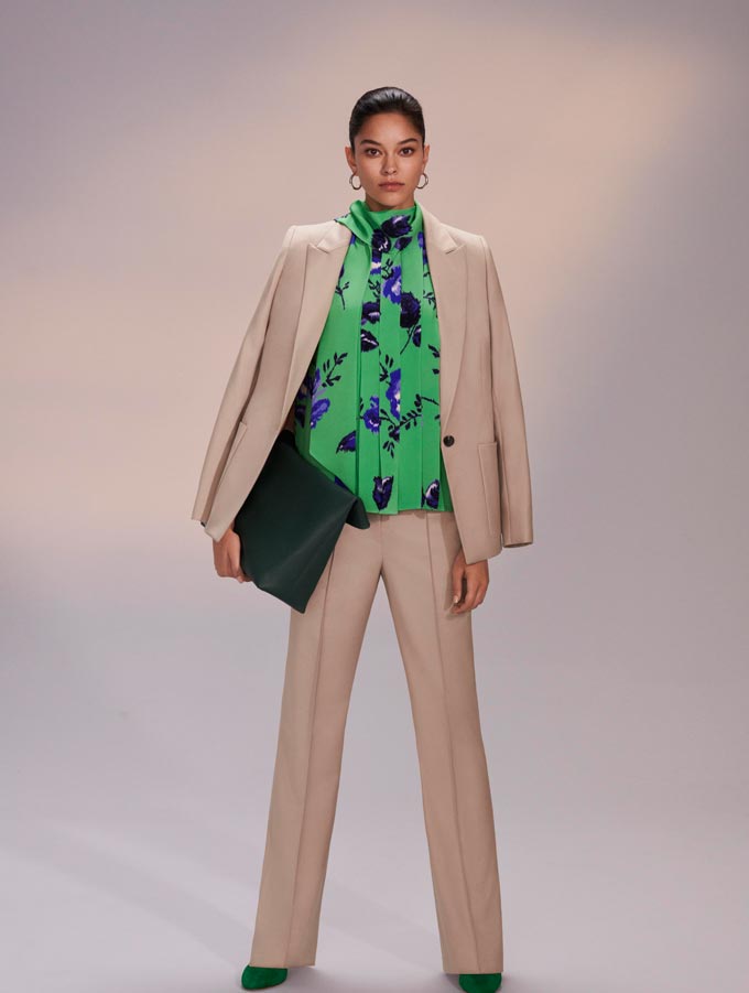 Women's power suits. A beige power suit paired with a green top with a blue flower print and an oversized black clutch. Image: Hobbs.