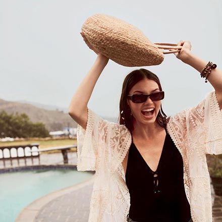 A woman in a black one piece suit wearing golden hoop earrings and holding a straw bag over her head as she's walking by a pool. Image: Accessorize.