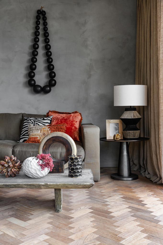 Partial view of a living space with a herringbone pattern parquet flooring, a grey sofa, a reclaimed wood bench as a coffee table and eclectic decor. Image: Amara.