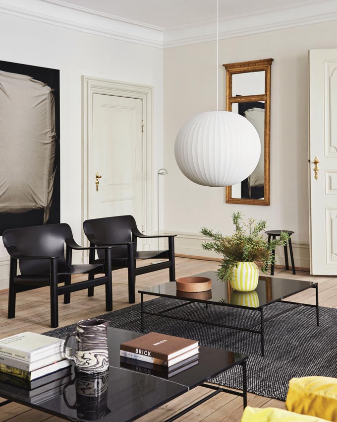 A contemporary living room featuring a white pendant light hanging over two glasstop coffee tables and two black Hay Bernard lounge chairs with a Bauhaus flair. Via Nest.co.uk.