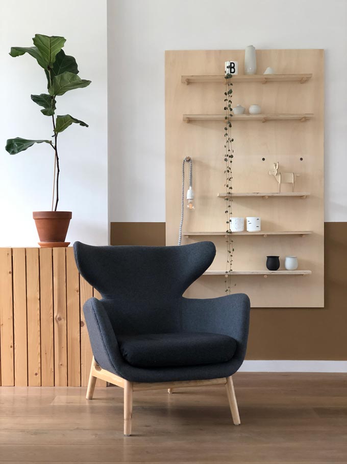 A statement winged armchair in a dark grey fabric upholstery against a rack of shelves. Via Cult Furniture.