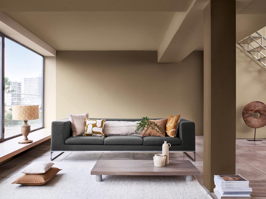 A contemporary living space with large windows, a dark grey sofa, a low coffee table and walls in Dulux Brave Ground beige. Via Dulux.