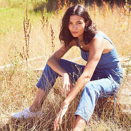 A woman outdoors in a grain field wearing a casual denim overalls outfit paired with white sneakers. Via Monsoon.